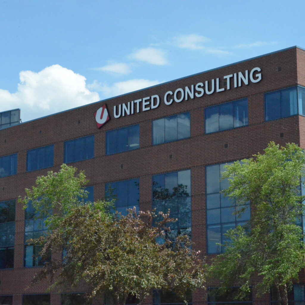 Brick office building with a 'UNITED CONSULTING' sign on the roof, glass windows, and green trees in front.